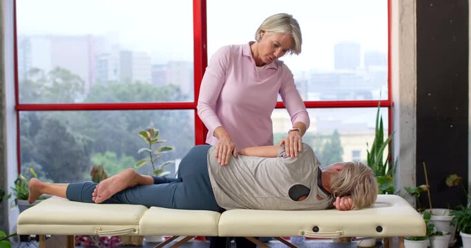 Senior therapist placing hands on patient, mobilizing spine on clinic table by window for mobility