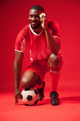African male kneeling in studio wearing red soccer kit holding gold medal and touching soccer ball © wavebreak3