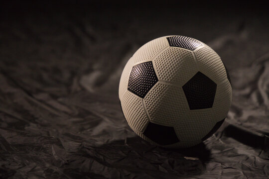 Textured black and white soccer ball is resting on dark crinkled drape, lit from upper left
