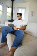Fototapeta premium African American man sitting on beige sofa at home holding tablet beside wood-burning stove