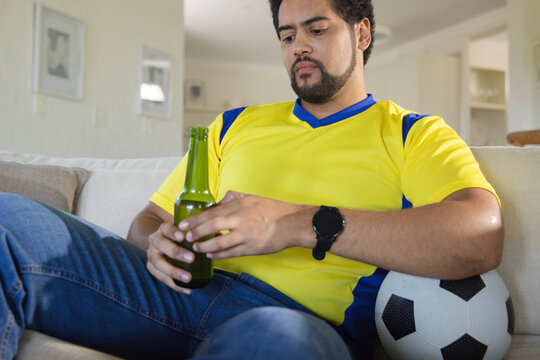 African American man sitting in living room in soccer jersey holding beer bottle beside soccer ball