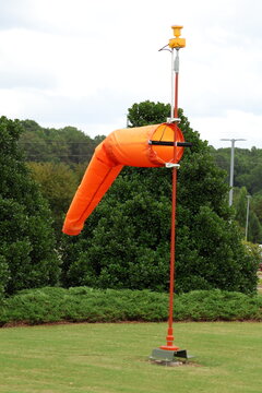 Bright orange windsock mounted on a tall pole against trees and cloudy sky background.