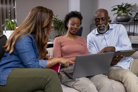 Diverse team leaning in pointing at silver laptop, holding tablet, pink notebook in office lounge