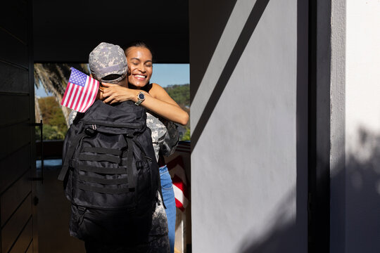 African American woman hugging soldier in camo at doorway with tactical pack and small US flag