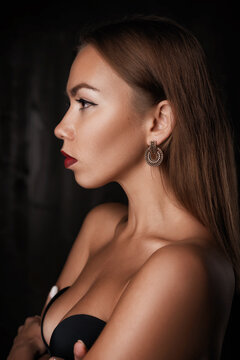 Side profile of a young caucasian woman with long straight hair, a gold hoop earring and red lipstick, bare shoulders and a black strapless bra in low light.
