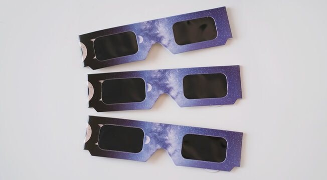 Close-up flat lay view of protective solar eclipse paper glasses featuring a cosmic purple night sky and moon phases design, arranged neatly on a white surface for astronomy and space event concepts.