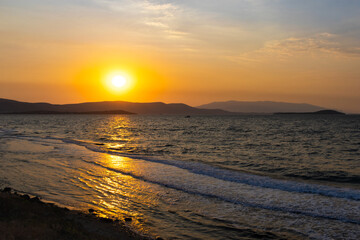Obraz premium Breathtaking golden sunset over a gently rippling sea with rolling waves and foamy shore, backed by dark silhouettes of distant hills and a hazy island horizon.