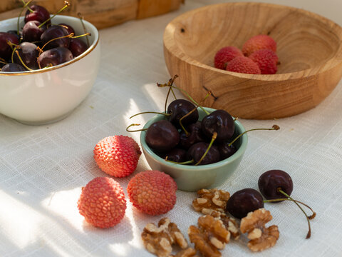 Dark sweet cherries, Prunus avium, lychee fruits, Litchi chinensis, and walnut kernels, Juglans regia, arranged in ceramic and wooden bowls on white linen. Rustic natural food composition featuring fr