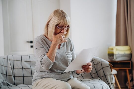 Senior woman with blonde hair wearing glasses sits on a couch, holding a document and looking thoughtfully at the paper in a well-lit living room