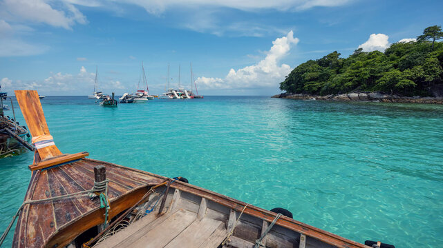 View of a traditional wooden long-tail boat floating on clear turquoise water with sailboats anchored near the lush coastline of Ko Racha Yai, Phuket, Phuket, Thailand.