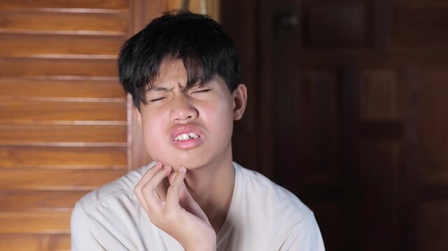Close-up of a teenage boy grimacing in pain, touching his swollen cheek after wisdom tooth extraction surgery, expressing discomfort from dental pain.	