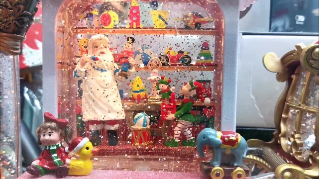 Christmas snow globe with Santa Claus, elves and colorful toy figurines on shelves. Falling snow and festive decoration display for winter holiday celebration and childhood wonder concept