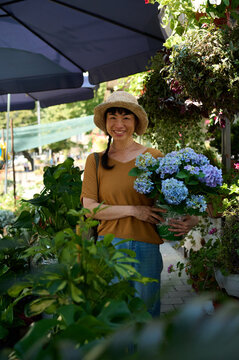 Smiling Asian Woman With Hydrangea Flowers At Outdoor Plant Market Wearing Straw Hat And Casual Clothes