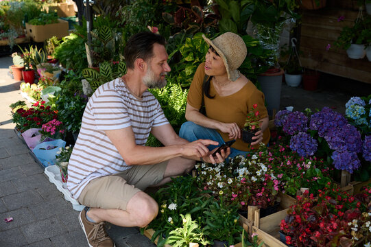 Couple Shopping Plants at Outdoor Plant Market &mdash; Man Showing Phone to Asian Woman in Hat