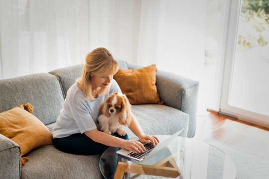 Smiling blond haired woman with her puppy sitting at home and using a laptop