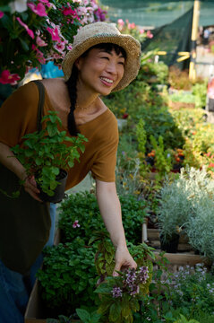 Asian Woman Smiling and Selecting Potted Herbs at Outdoor Plant Nursery Market in Straw Hat