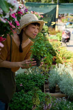 Asian Woman Shopping for Potted Herbs at Outdoor Garden Center Wearing Straw Hat and Casual Clothes