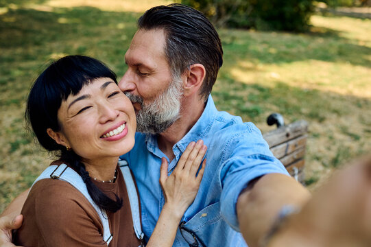 Bearded Man Kissing Asian Woman on Park Bench While Taking a Selfie &mdash; Loving Couple in Summer