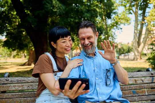 Smiling Asian Woman and Bearded Man Video Calling on Park Bench While Waving During Summer