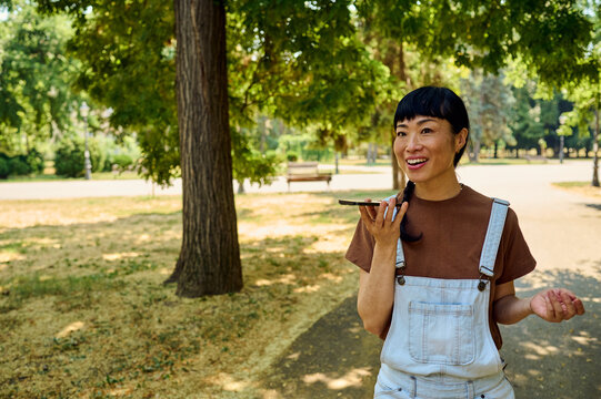 Asian Woman Speaking Into Smartphone While Smiling In Sunny Park During Casual Outdoor Conversation