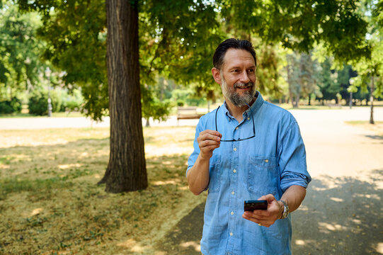 Smiling Middle-Aged Man Walking in Park Holding Smartphone and Glasses in Casual Denim Shirt