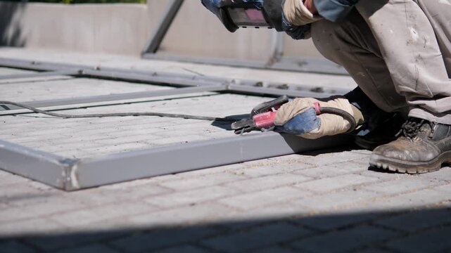 Worker remains crouched on gray rooftop while using handheld power tool beside metal framing, with thick cable lines, protective gloves, rugged boots, and soft focused industrial background in