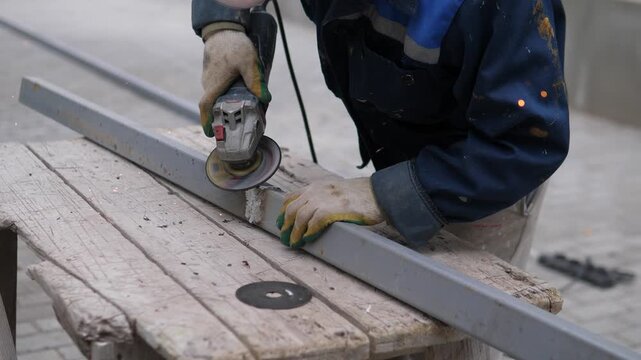 A worker is grinding metal in a focused and industrious workshop setting, with blue and gray tones. The close-up side view highlights the angle grinder and metal beam, emphasizing.