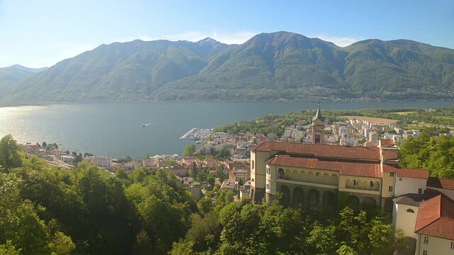Panoramic view above Sanctuary of Madonna del Sasso in Locarno, scenic Lake Maggiore landscape with calm water reflections, mountains and distant boat on sunny summer day in Switzerland