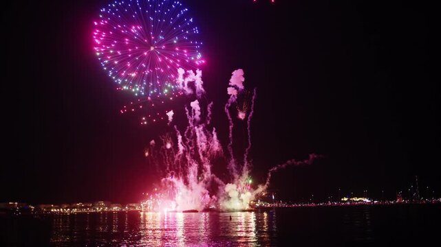 Stunning nighttime celebration with vibrant fireworks exploding over calm ocean water, reflecting colorful lights.