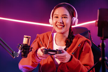 Host channel of smiling beautiful Asian girl streamer with joystick playing online game wearing headphones paste talking with viewers media online. Esport skilled team players in neon room. Stratagem. © InfiniteFlow