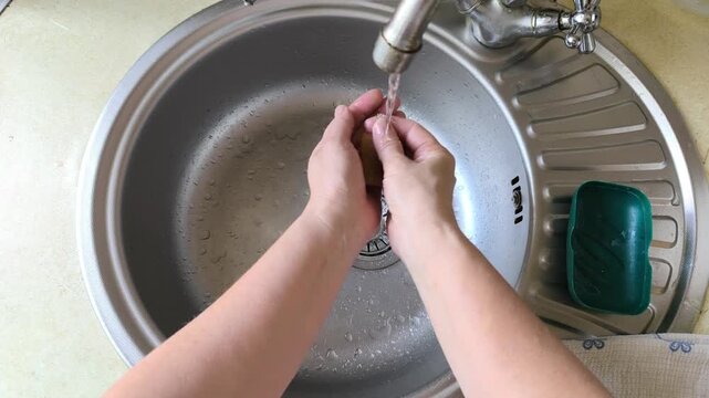 Female hands washing with soap bar under running water in kitchen sink. Hand rubbing brown soap for hygiene in silver sink bowl. Domestic process involves washing hands for health at home.