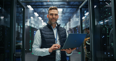 Fototapeta premium Portrait of Network Engineer Holds Laptop Computer in Data Center Corridor, Performing Server Health Checks, Updating Firmware, Troubleshooting Data Storage Systems and Documenting Rack Inventory.