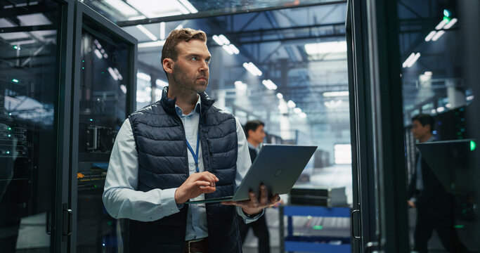 Male IT Specialist Uses Laptop Computer for Server Maintenance and Diagnostics While Inspecting Rack Hardware, Performing Network Performance Checks and Equipment Audit in a Data Center Facility.