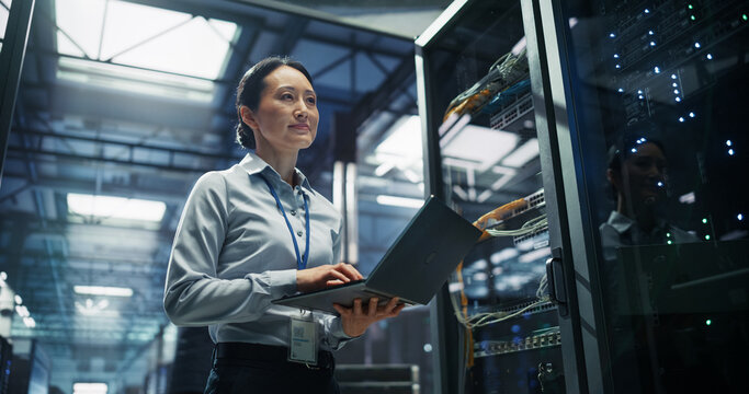 Asian Female It Support Engineer Holds Laptop Computer at Open Network Cabinet in Data Center, Auditing Ethernet Patch Cables, Testing Bandwidth Throughput and Deploying Cloud Server Updates.