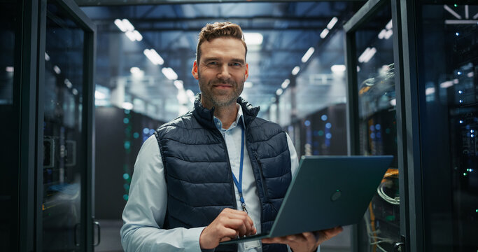 Portrait of Data Center Technician Uses Laptop Computer Between Server Racks, Running System Maintenance and Diagnostics, Monitoring Data Storage Operations and Managing Web Hosting Infrastructure.