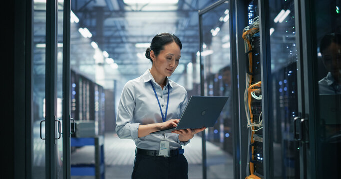 Asian Female System Administrator Using Laptop Computer for Software Configuration and Data Backup Monitoring While Standing Between Rows of Server Cabinets in AI Cloud Computing Center.