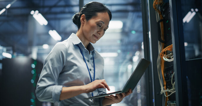 Asian Female It Specialist Holding Laptop Computer and Working on Firmware Updates and Server Performance Checks Next to Rack Cabinet With Network Cabling in an AI Cloud Computing Data Center Facility