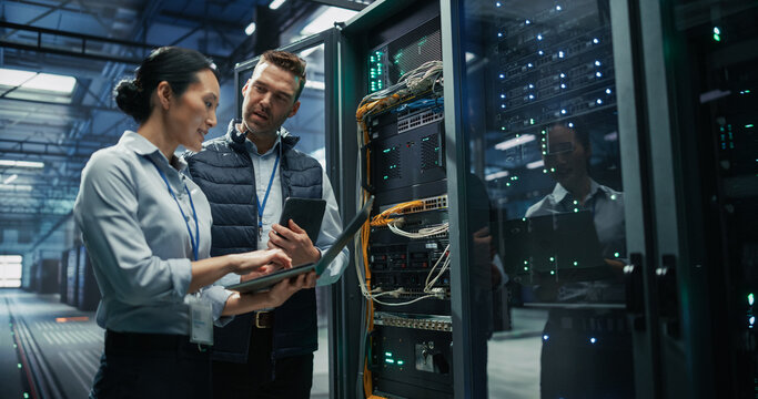 Asian Female Software Developer and Caucasian Male Maintenance Engineer Working on Laptop and Digital Tablet Computer to Plan Data Backup and System Beside Open Server Rack Cabinet in AI Data Center.