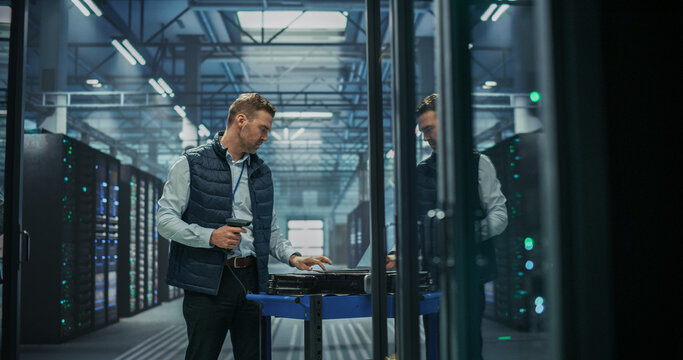 Caucasian Male It Specialist Working on Laptop Computer While Holding Barcode Scanner Beside Maintenance Cart With Server Units to Track Hardware Assets and Log Equipment Data in AI Data Center.