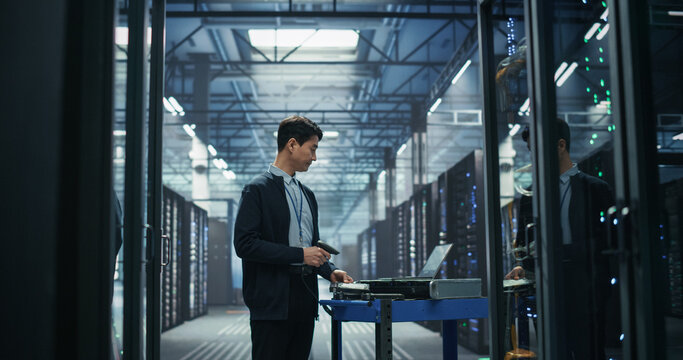 Asian Male Network Technician Uses Handheld Barcode Reader to Register New Server Hardware on Mobile Cart Beside Laptop, Performing Inventory Audit and Equipment Provisioning in AI Data Center.