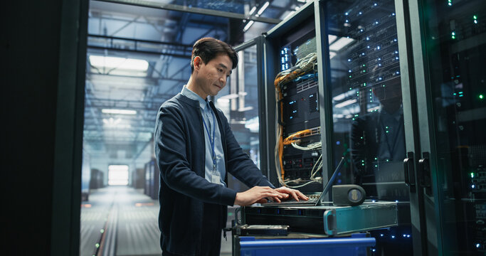 Asian Male Systems Engineer Using Laptop Computer on Mobile Workstation Cart Beside Opened Server Rack While Performing Software Updates and Network Diagnostics in AI Data Center