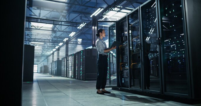 Full Length Shot of Female Asian Data Center Engineer Holding Laptop Computer While Inspecting Open Server Rack Cabinet, Performing Hardware Configuration and Network Maintenance in AI Facility