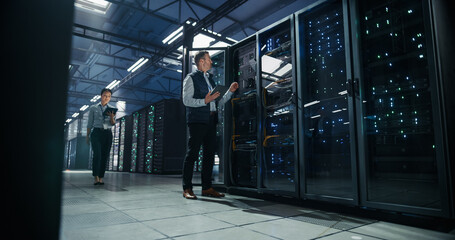 Caucasian Male Engineer Uses Tablet Computer to Inspect Open Server Rack Cabling While Asian Female...