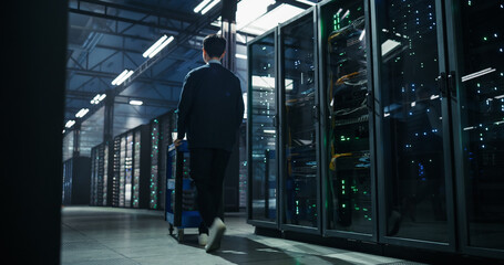 Rear View of Asian Male Engineer Pushing Equipment Cart Along Corridor of Server Racks While...