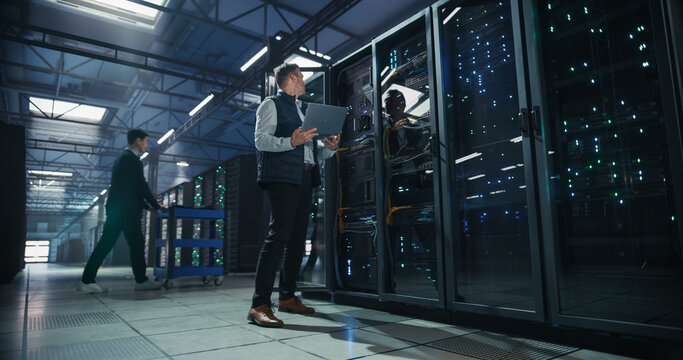 Two It Specialists at Work in AI Data Center: Caucasian Male Engineer With Laptop Computer Configuring Server Rack While Asian Colleague Transports Hardware on Cart Through Data Warehouse