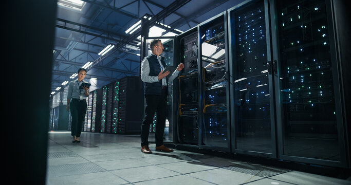 Caucasian Male Engineer Uses Tablet Computer to Inspect Open Server Rack Cabling While Asian Female Colleague Approaches With Device, Performing Hardware Diagnostics in AI Cloud Data Center
