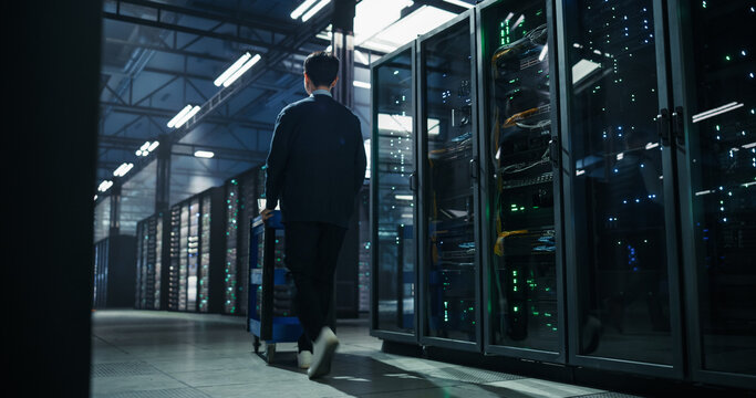 Rear View of Asian Male Engineer Pushing Equipment Cart Along Corridor of Server Racks While Transporting Hardware for Installation and Upgrade in AI Cloud Computing Data Center Facility