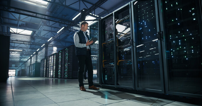 Wide Angle Shot of Caucasian Male System Administrator Standing Beside Open Server Rack Cabinet and Using Tablet Computer for Network Monitoring and Maintenance in Modern AI Cloud Data Center