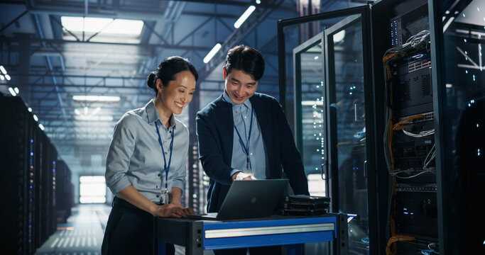 Two Asian It Specialists, Man and Woman, Discuss Server Configuration on Laptop Computer Placed on Maintenance Cart With Hard Drives Next to Open Rack Cabinet in AI Cloud Computing Data Center