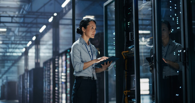 Asian Female System Administrator Working With Tablet Computer Beside Server Rack Cabinet, Performing Firmware Update and Data Storage Audit in High Tech AI Cloud Computing Data Center Facility